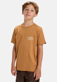Camiseta de algodón marrón con mangas cortas y cuello redondo; presenta una ligera impresión del logo en el pecho y una pequeña etiqueta negra en la manga.