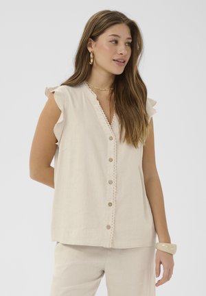 Naine, kellel on pikad pruunid juuksed, kannab varrukateta beežikat nööbitavat pluusi rüüsidega õlgadel ja sobivaid pükse, aksessuaaridena ehted.