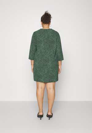 Femme de dos, portant une robe verte à hauteur du genou avec un motif de feuilles noires et des sandales à talons noires, sur fond uni.