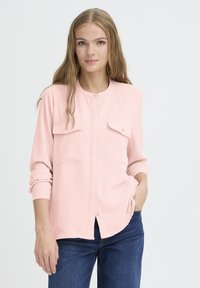 Chemise boutonnière rose clair à manches longues, avec deux poches poitrine et un col rond, associée à un jean en denim. Texture de tissu lisse.