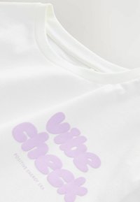 T-shirt bianco con una grande grafica testuale di colore lilla chiaro che recita "Vibe Check" e un sottotitolo "Positive Energy Era" sottostante. Tessuto morbido.