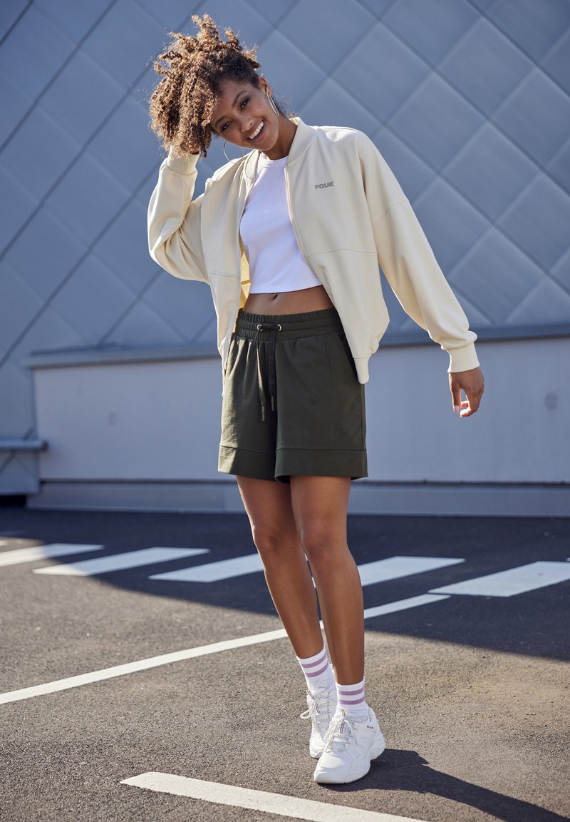 Light cream zip-up jacket over a white crop top, paired with green shorts and white sneakers. The model stands on a paved surface.