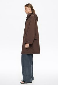 Cazadora ligera marrón con capucha, bolsillos laterales y botones de presión. Combinada con pantalones anchos de color azul oscuro que presentan detalles de costura.