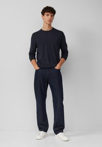 Marineblauwe lange mouwen crew neck shirt, gecombineerd met losse donkere spijkerbroeken en witte sneakers, tegen een neutrale achtergrond.