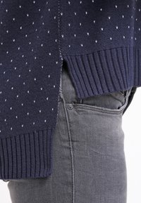 Marinblå tröja med vitt diagonalt mönster, ribbad nederkant och sprund i sidorna. Bärs över grå jeans, vilket framhäver den texturerade tyget och den avslappnade passformen.
