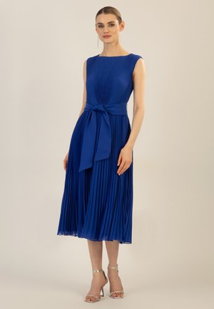 Pleated blue dress with a fitted bodice, cap sleeves, and a matching waist tie. The skirt is flowy, reaching mid-calf. Silver heeled sandals.