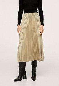 Jupe midi plissée beige en satin, avec un design fluide et une taille structurée, associée à un haut noir ajusté et des bottes jusqu'au genou.