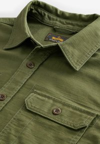 Olive green shirt with a pointed collar, chest pocket, and four brown buttons. Made of durable fabric with visible stitching details.