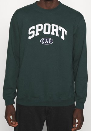 Ciemnozielona bluza z białym napisem „SPORT” nad owalem z napisem „GAP”, noszona przez osobę o ciemnej karnacji i czarnych spodniach.