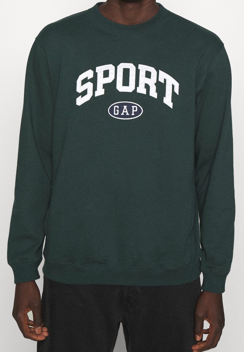 Sweat-shirt vert foncé avec le texte blanc "SPORT" au-dessus d'un ovale contenant "GAP", porté par une personne à la peau foncée et portant un pantalon noir.