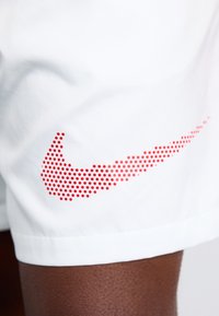 Bijele atletske kratke hlače s velikim crvenim točkastim Nike swoosh logotipom sa strane. Lagana tkanina s glatkom teksturom.