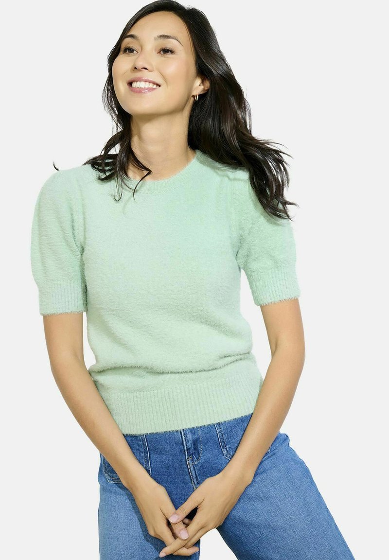 Šviesiai žalias, trumpomis rankovėmis megztinis su tekstūrinėmis, pūkuotomis mezgimo medžiagomis. Raukšlėtos rankogaliai ir apačia, derinamas su mėlynais džinsais.