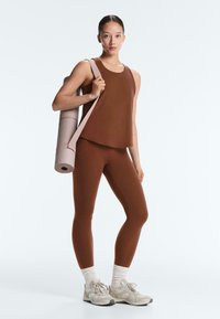 Fiatal nő barna sportos ruhában, vállán egy összehajtott jógaszőnyeggel, fehér zoknit és tornacipőt viselve, fehér háttér előtt.