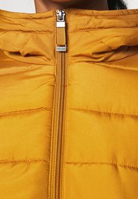 Gelbe gepolsterte Jacke mit einem Reißverschluss, die eine Kapuze und eine gesteppten Textur aufweist. Reißverschlusszüge und Markenlabel sind sichtbar.