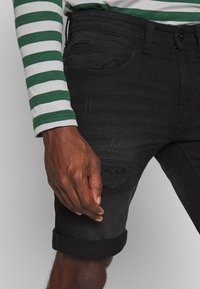 Pantaloni corti in denim neri con risvolti, caratterizzati da una toppa strappata e sottili righe verticali. Indossati con una camicia a maniche lunghe a righe verdi e bianche.