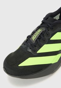 Zwarte sportieve schoen met groene accenten, voorzien van een textuurstof bovenwerk, veters en het "ADIZERO"-logo op de tong.
