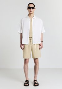 Uomo che indossa occhiali da sole neri, camicia bianca a maniche corte con bottoni sopra una canotta beige, pantaloni corti beige con cintura, sandali neri, in piedi contro uno sfondo uniforme.