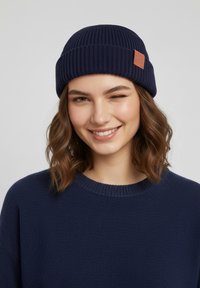 Navy ribbed knit beanie with a leather patch accent. The model wears a matching navy sweater with a round neckline.