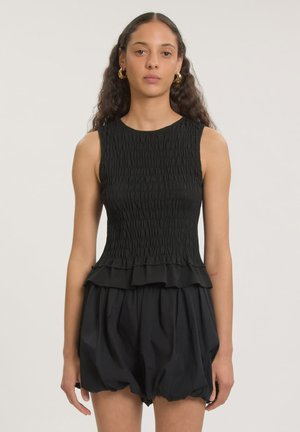 Jonge vrouw met lang krullend haar draagt een mouwloze zwarte top met textuur en losse zwarte shorts tegen een effen lichte achtergrond.
