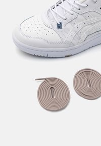 Vit lädersneaker med perforeringar, blå accenter och en texturerad sula. Inkluderar två rullade, beige snören med spetsiga ändar.