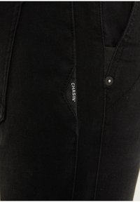 Pantalones de mezclilla negros con un diseño estilizado que presenta una pequeña etiqueta y un detalle de botón cerca del bolsillo. Tejido texturizado con costuras sutiles.