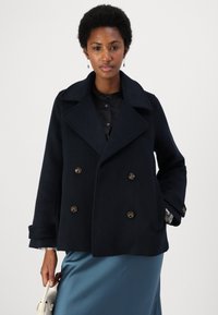 Marineblå dobbeltspent ullfrakk med stor utskjært krage, gullfargede knapper og sidelommer. Modellen har den på seg sammen med et skinnskjørt i blå sateng.
