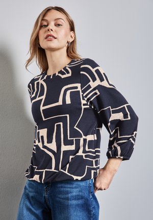 Marineblauw top met abstract beige patroon, ronde hals en driekwart mouwen. Zachte stof met subtiele textuur. Gecombineerd met blauwe jeans.