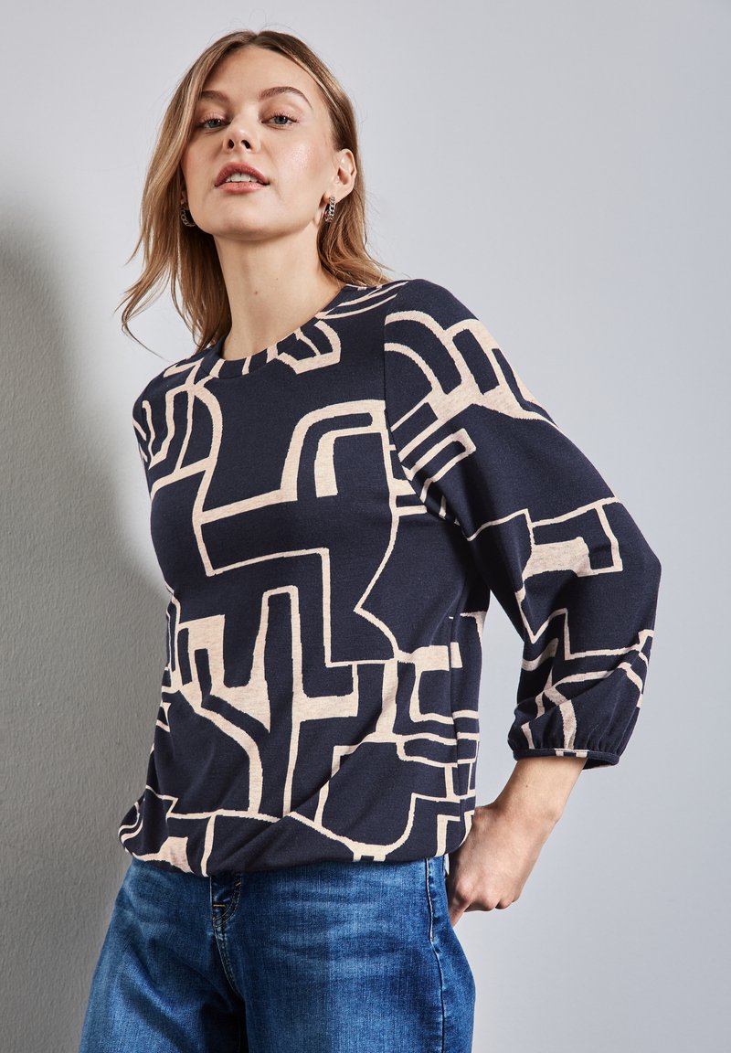 Top bleu marine avec un motif abstrait beige, encolure ronde et manches trois-quarts. Tissu doux avec une texture subtile. Assorti à un jean bleu.