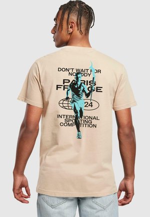 Beige cotton t-shirt featuring a graphic design of a runner with a torch. Text includes "PARIS 2024" and "INTERNATIONAL SPORTING COMPETITION."