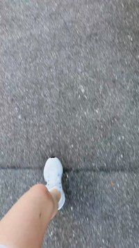 Weißer Sportschuh an einem Bein, das über einen strukturierten grauen Bürgersteig läuft. Der Schuh hat Schnürsenkel und ein rundes Zehendesign.
