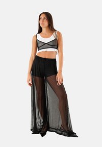 Pantaloni neri con sovrapposizione trasparente abbinati a un crop top bianco, con una sezione in rete nera. L'outfit mette in evidenza il tessuto fluido e il design aderente.