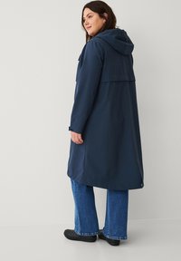 Marineblauwe waterdichte jas met capuchon, voorzien van een rugdeel, lange mouwen en verstelbare manchetten, gecombineerd met uitlopende spijkerbroeken en zwarte schoenen.