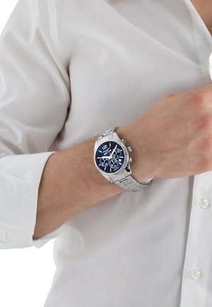 Orologio in acciaio inossidabile argentato con quadrante blu, dotato di sottoquadranti cronografici e una lunetta texturizzata. Indossato al polso, con una camicia bianca visibile.