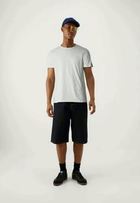 T-shirt branca de mangas curtas, calções de ganga escuros, sapatilhas pretas, boné azul e óculos de sol amarelos. Conjunto casual numa pose de pé.