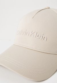 Beige caps laget av teksturert stoff med buet skjerm; har hevet Calvin Klein-logo i matchende farge.