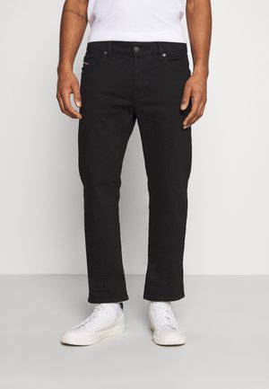 Mann trägt schwarze Jeans mit geradem Bein, weiße Sneaker und ein weißes Hemd, steht vor einem schlichten hellen Hintergrund.