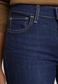 Plano detalle de unos vaqueros de mezclilla azul oscuro con costuras amarillas visibles y un bolsillo frontal, junto a una persona que lleva una camiseta de manga larga marrón.