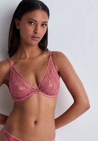 Soutien-gorge en dentelle de couleur rose doux avec des panneaux transparents, des bretelles réglables et une bande inférieure lisse ornée d'un accent en métal doré au centre.