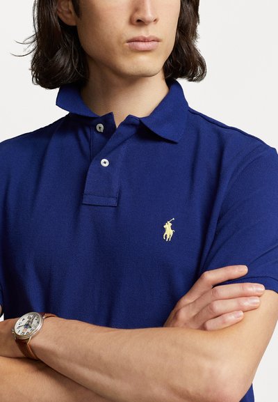Tumesinine polo särk, mis on valmistatud tekstuuriga kangast; sisaldab klassikalist kraed, kahte nuppu ja väikest kollast logo rinnal. Nähtav pruun kell.