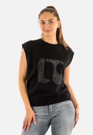 Maglione nero senza maniche con colletto e orlo a coste, caratterizzato da un logo argentato scintillante sul davanti. Indossato con jeans grigio chiaro.