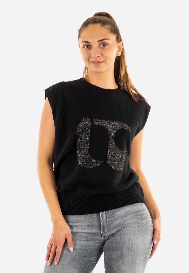 Suéter negro sin mangas con un escote y dobladillo acanalados, que presenta un logo plateado brillante en la parte delantera. Llevado con jeans gris claro.