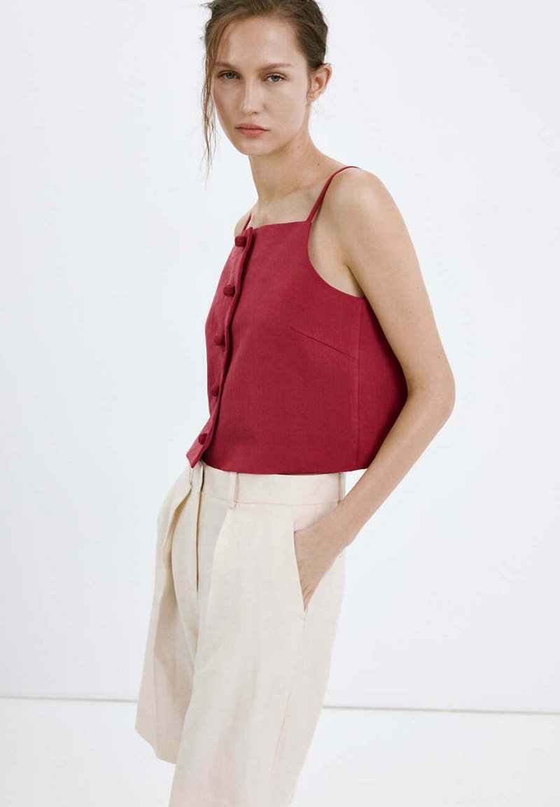 Femme portant un haut rouge sans manches boutonné et un pantalon beige taille haute à jambes larges, une main dans la poche, debout devant un fond blanc.