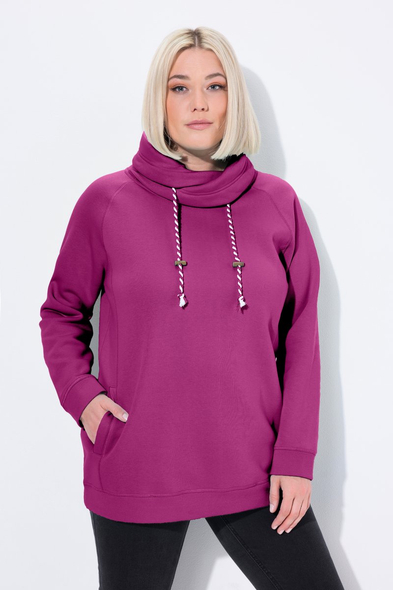 Vrouw met kort blond haar die een magenta pullover-hoodie draagt met gestreepte trekkoorden en zwarte broek, staand tegen een witte achtergrond.