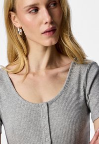 Kvinde med langt blondt hår iført sølvfarvede øreringe og en grå ribstrikket top med knapper, kigger mod venstre på en ensfarvet baggrund.