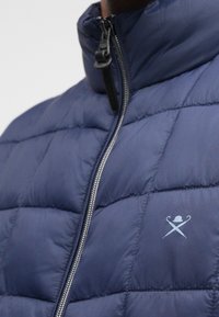 Ναυτική επενδεδυμένη παλτό με ψηλό γιακά, κλείσιμο με φερμουάρ και λογότυπο στο στήθος. Διαθέτει απαλή, γυαλιστερή υφή και κουτάκι ραφή.