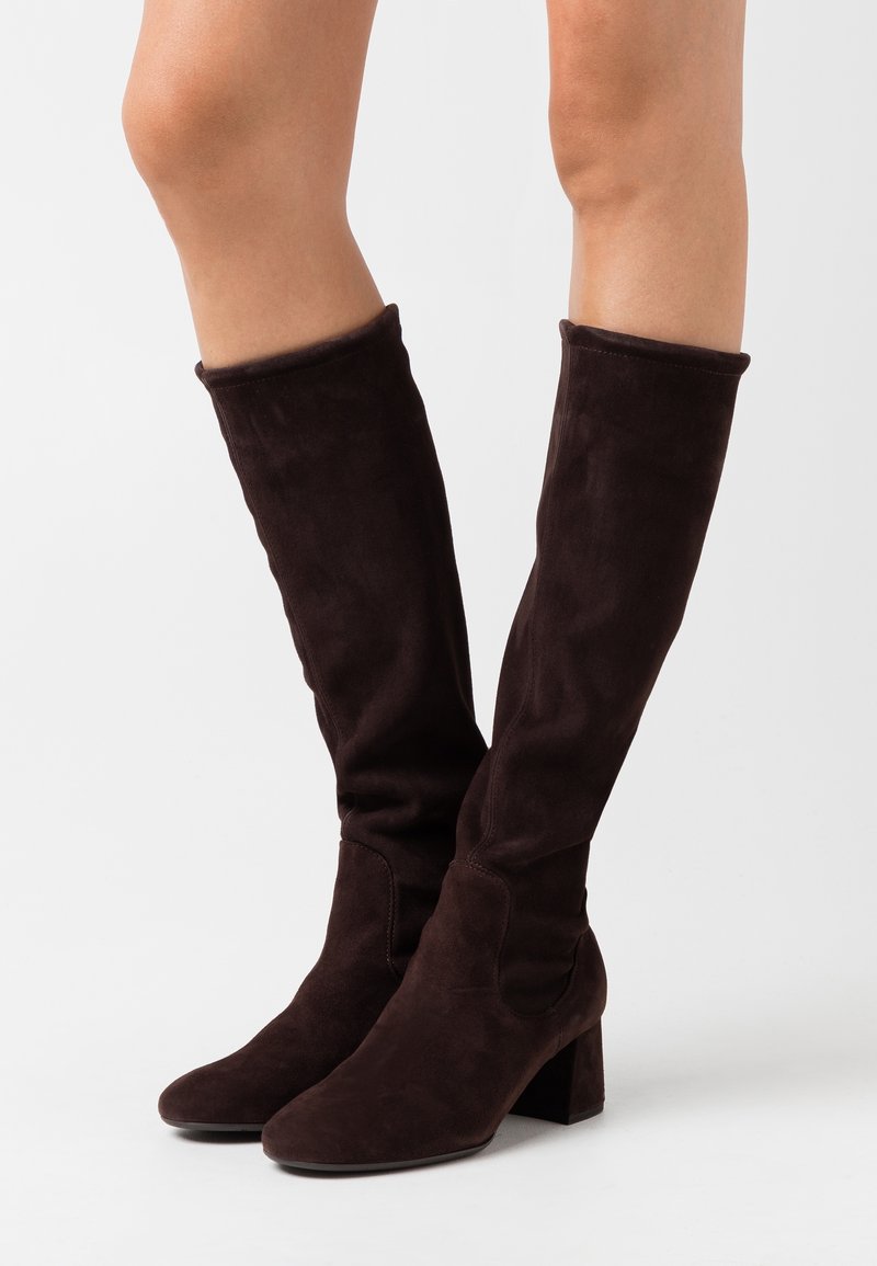 Bottes hauteur genou en daim marron foncé à talons carrés, portées sur des jambes nues sur un fond clair uni.