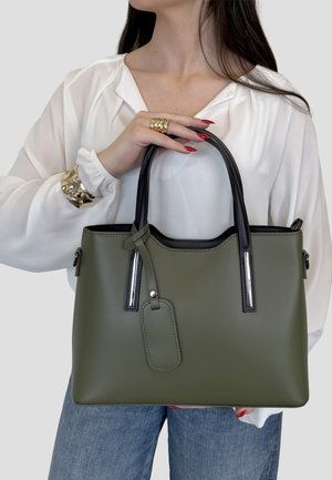 Vrouw die een olijfgroene lederen shopper met zwarte handvatten vasthoudt, gekleed in een wit blouse, blauwe jeans, gouden armband en ringen, met rode nagels.