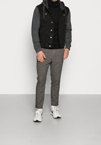 Schwarze Denim-Weste mit grauen Ärmeln, über einem schwarzen Rollkragenpullover getragen. Graue karierte tapered Hose und weiße Sneakers mit schwarzen Akzenten.
