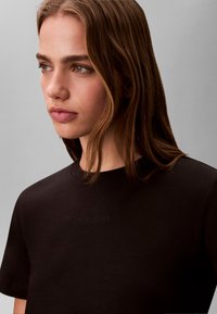 Young woman with brown hair wearing a black Calvin Klein T-shirt, looking slightly to the left against a plain gray background.