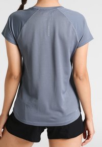 Camiseta gris de rendimiento con mangas cortas, tejido texturizado y dobladillo curvado. Presenta un logo sutil en la parte trasera. Combinada con pantalones cortos negros.